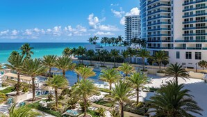 A resort with a pool area surrounded by palm trees, a beach, and a tall building with balconies. - 1BD/1.5BTH BeachFront Resort Style. Gym, WiFi, Free Parking, Jacuzzi, GameRoom. (Hollywood)