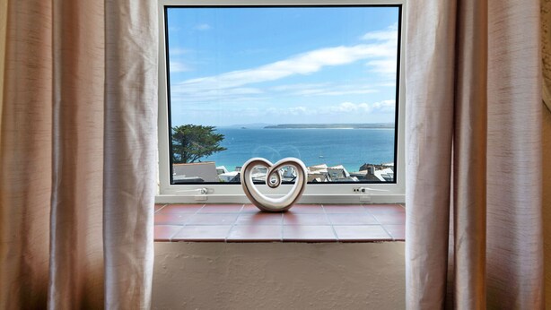 A view through a window with a heart-shaped object on the sill, overlooking a coastal area with buildings and trees.