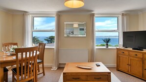 A room with a dining table, chairs, a TV, and a view of the sea.