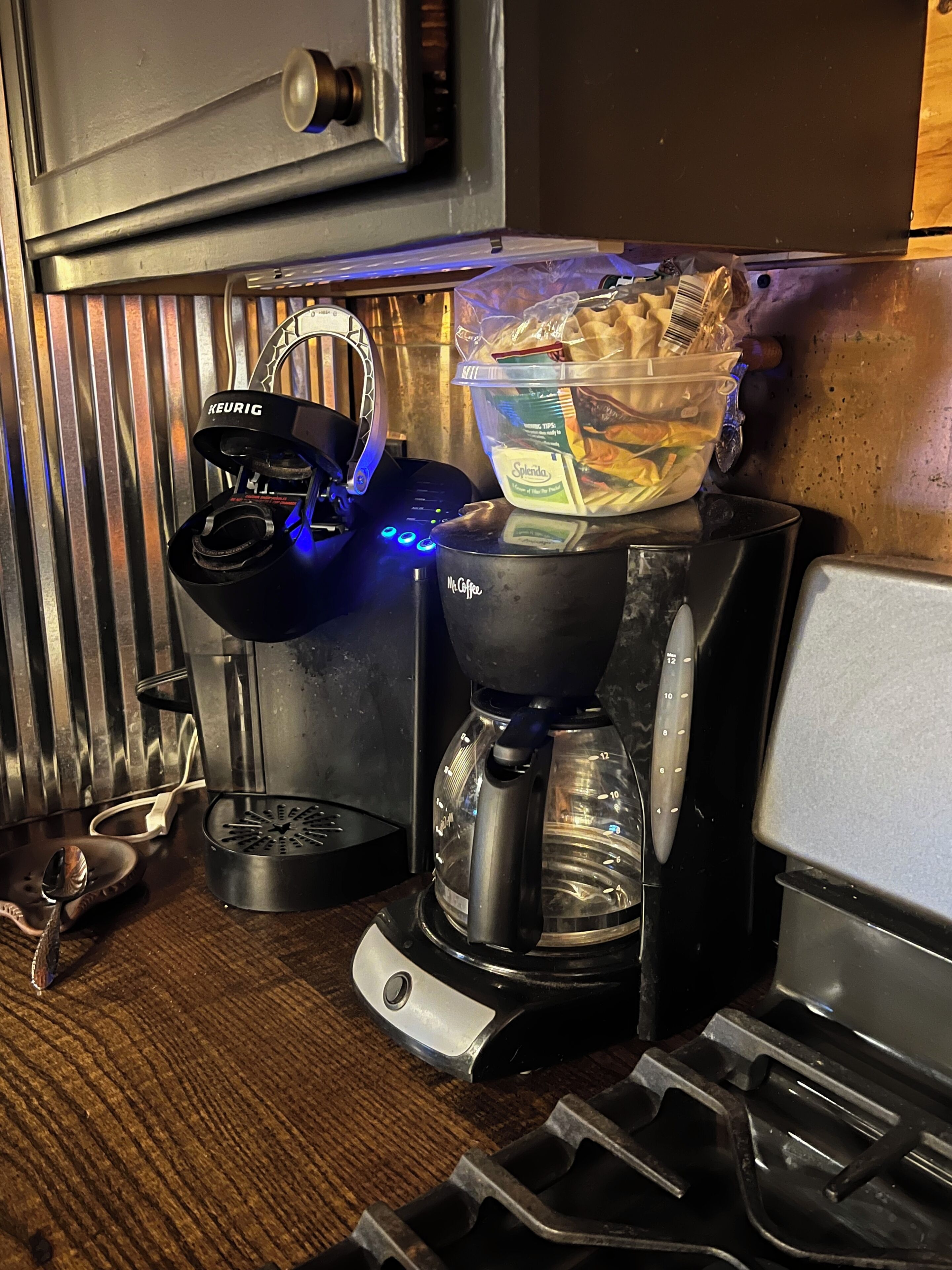 Una cafetera con un dispensador de cápsulas Keurig, una jarra de café Mr. Coffee y un recipiente con alimentos sobre una superficie de madera.