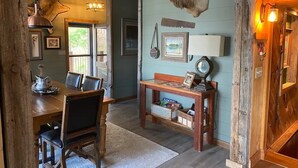 A rustic dining area with a wooden table and chairs, a hanging deer head, and a small side table with a lamp and picture frame.