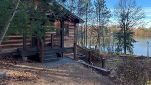 A log cabin with a wooden deck and stairs leading to a porch, surrounded by trees and a lake.