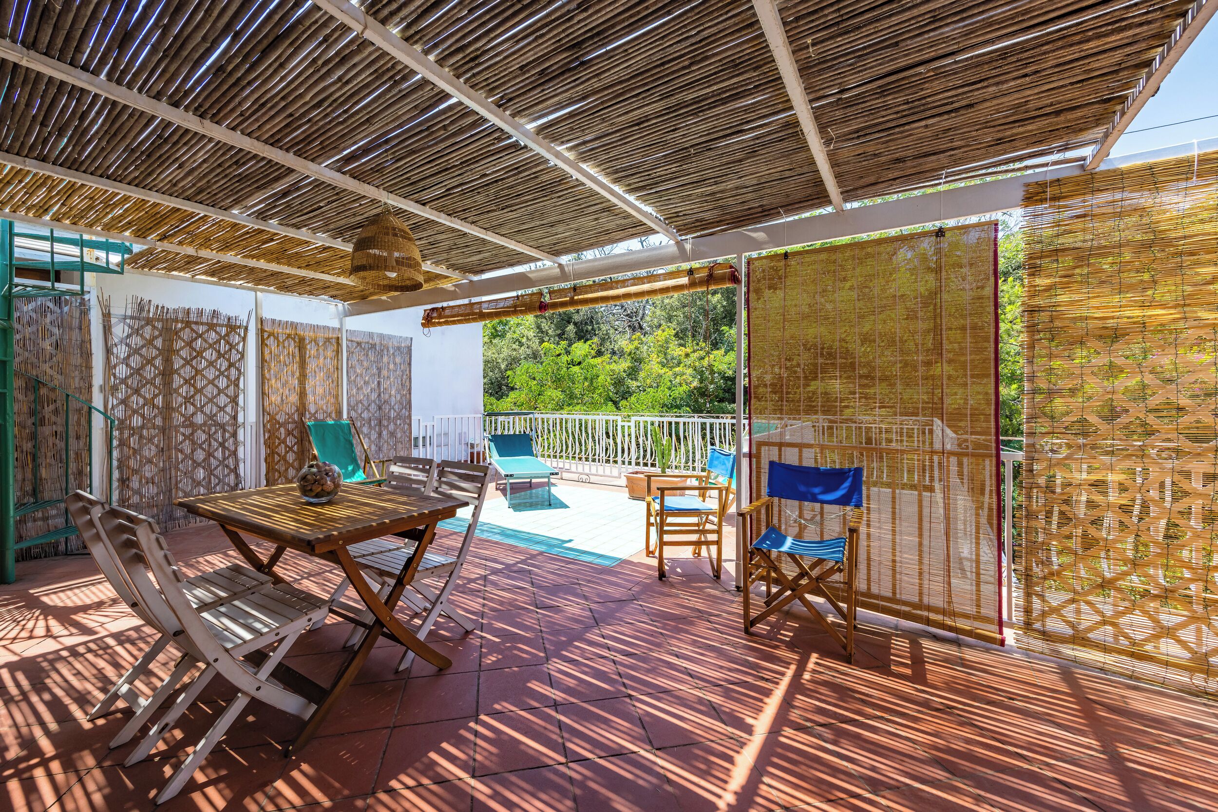 An outdoor patio area with a wooden table and chairs, a pool, and a view of greenery.