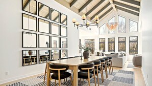 A dining area with a long table, multiple chairs, and a chandelier. There are large windows and a high ceiling with exposed beams. - Tremblant Prestige - Verbier 13-203 (Mont-Tremblant)