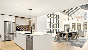 A modern kitchen with a central island, stainless steel appliances, and a dining area with a large window and a chandelier. - Tremblant Prestige - Verbier 13-203 (Mont-Tremblant)