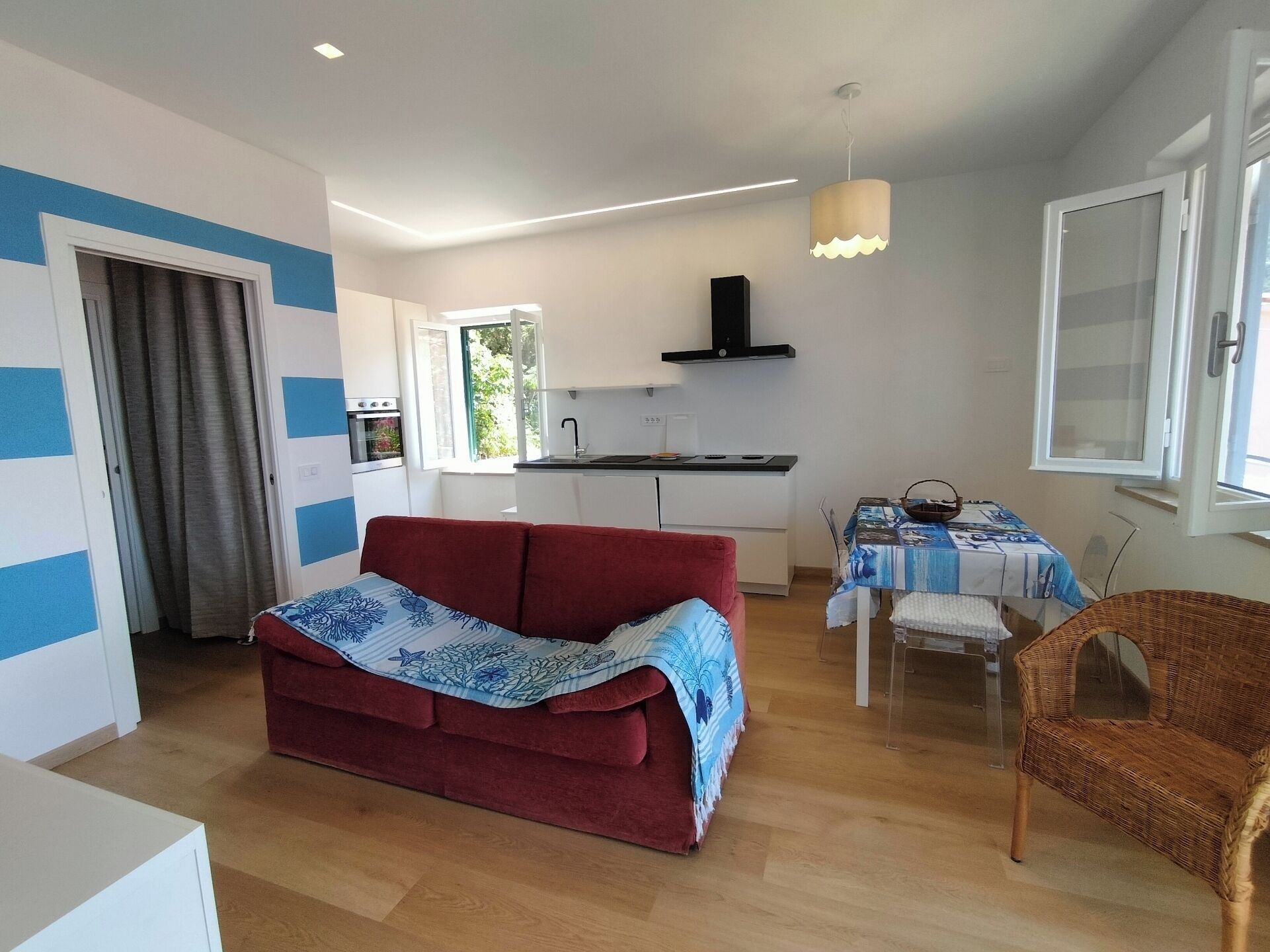 A compact living space with a kitchenette, a red sofa, a dining table with a blue tablecloth, and a wicker chair.