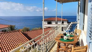 Un balcón con mesa y sillas, con vista al mar, techos de teja anaranjada y un cielo azul despejado.