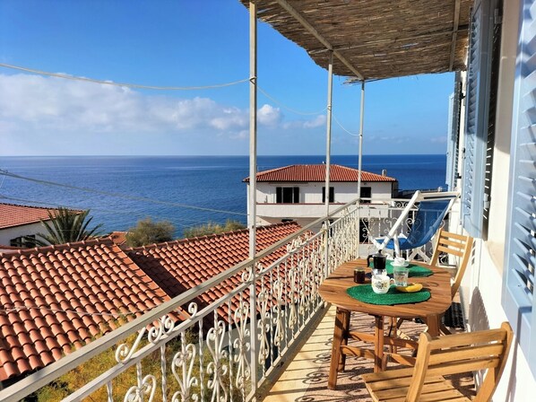 A balcony with a table and chairs, overlooking a coastal view with terracotta roofs and a clear blue sky.