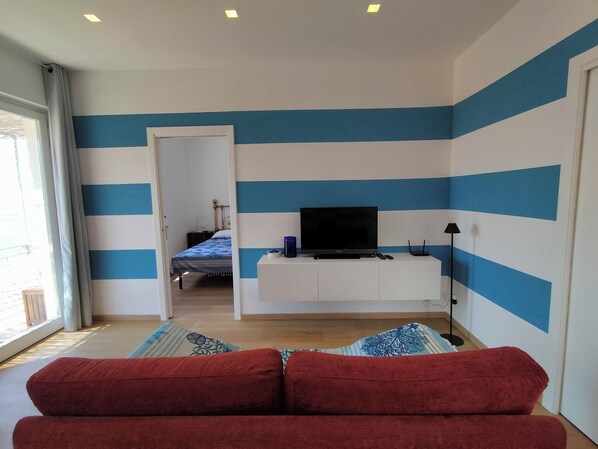 Una habitación con una televisión sobre un mueble blanco, un sofá rojo y una cama frente a una pared azul y blanca a rayas.