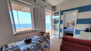 Una habitación con vistas al mar, una mesa con mantel, una silla roja y una cama frente a una pared azul y blanca a rayas.