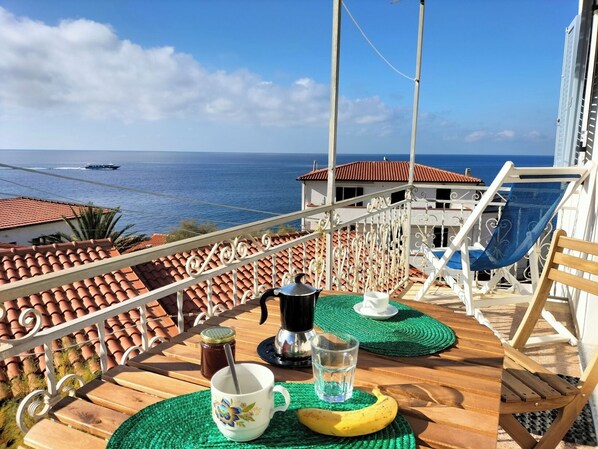 Un balcón con vista al mar, una mesa con una cafetera, una taza y un vaso de agua.