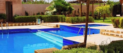 Une piscine dotée d’une structure au toit de chaume, entourée de haies soigneusement taillées et de palmiers.
