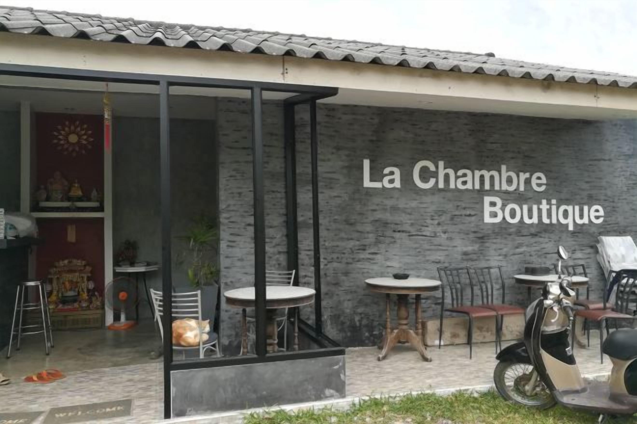 Fachada con el nombre 'La Chambre Boutique' en una pared de ladrillo. Hay áreas de estar al aire libre con mesas y sillas, y una motocicleta estacionada afuera.
