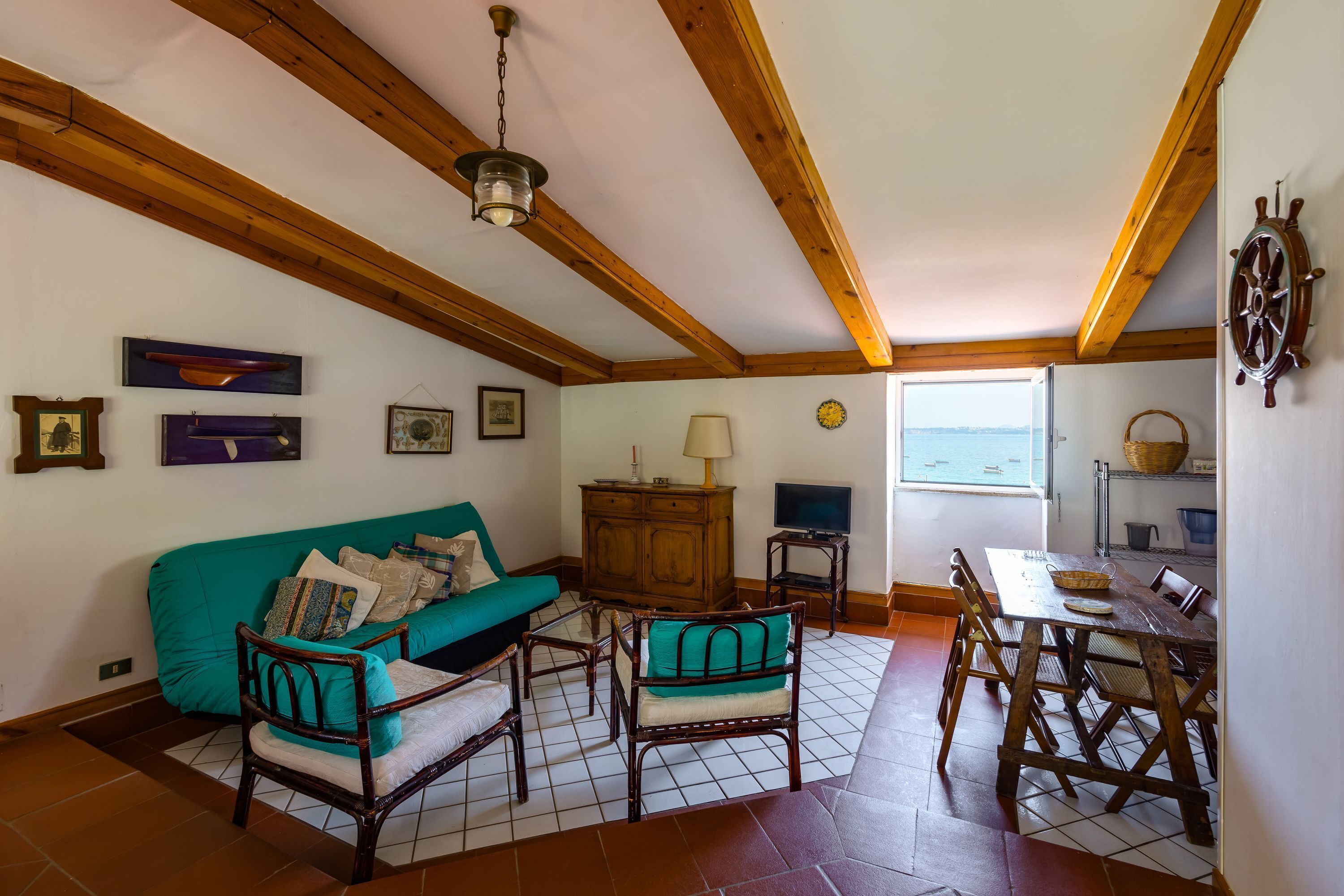 A cozy living room with a wooden ceiling, a teal sofa, wooden chairs, a dining table, and a television.