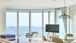 A modern, well-lit room with a large window, a flat-screen TV, and a glass dining table. - Ayia Napa Marina East Tower Suite (Ayia Napa)