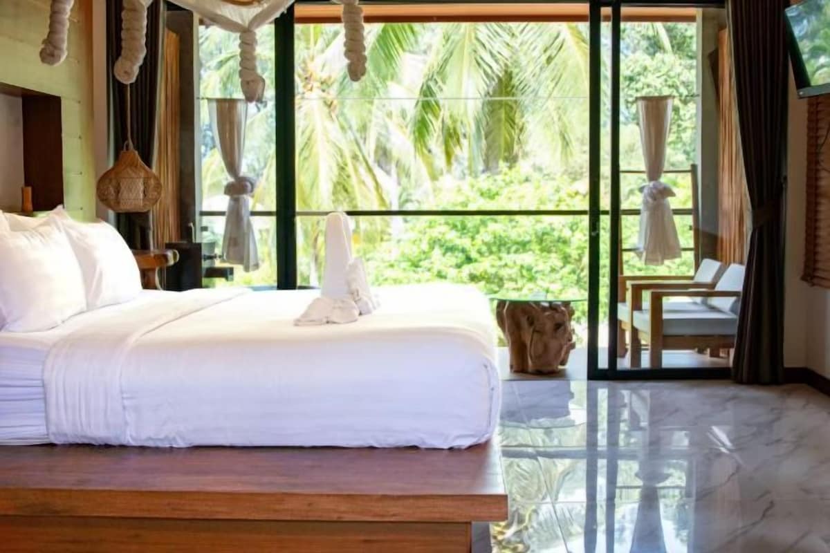 A bedroom with a large bed, a wooden headboard, a small table, and a view of greenery outside.