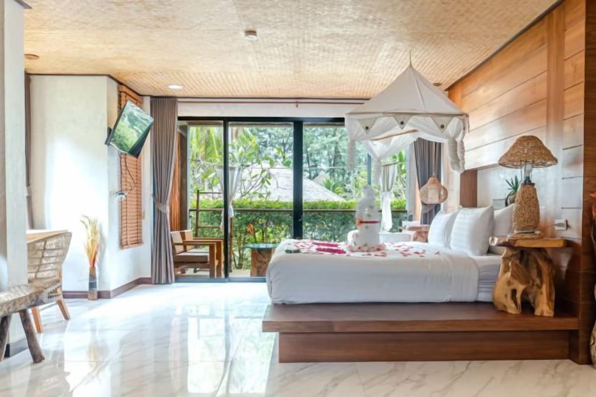 A bedroom with a large bed, a TV, a balcony with a table and chairs, and a view of greenery.