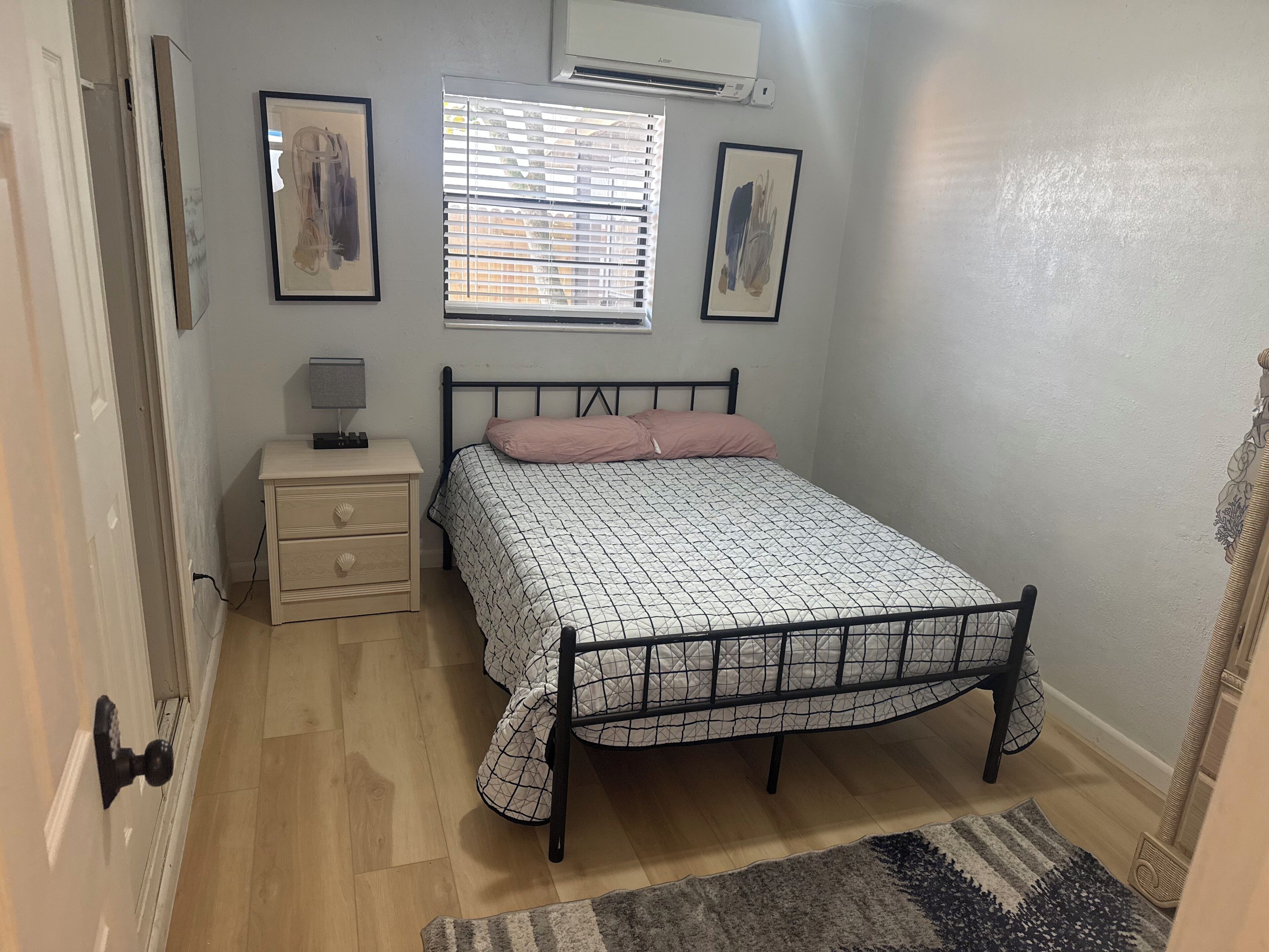 A bedroom with a bed, bedside table, lamp, air conditioning unit, and two framed pictures on the wall.
