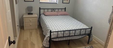 A bedroom with a bed, bedside table, lamp, air conditioning unit, and two framed pictures on the wall.