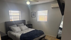 A bedroom with a bed, two windows with blinds, a bedside table, a ceiling fan, and a wall-mounted air conditioner. - Charming 3-bedroom house in wonderful Melbourne with WiFi, AC (Melbourne)