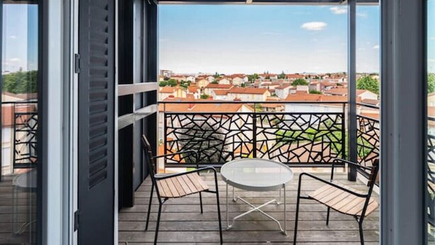 Un balcón con mesa y sillas, vista panorámica de la ciudad y barandilla metálica.