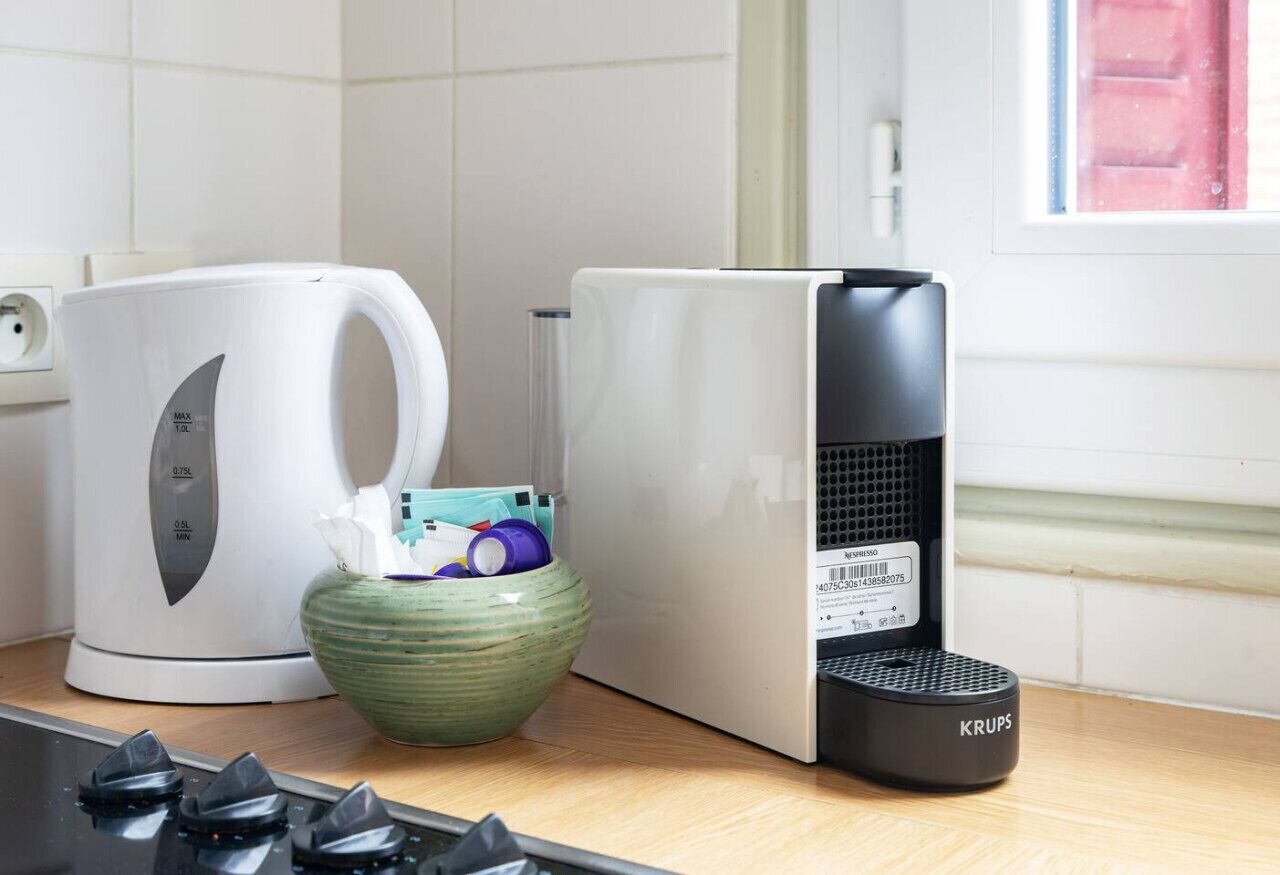 Une cuisine avec une bouilloire blanche, un saladier vert, une machine à café noire et une cuisinière.