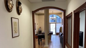 A hallway with a tiled floor, framed certificates on the wall, and a view into a room with a piano and a chandelier.