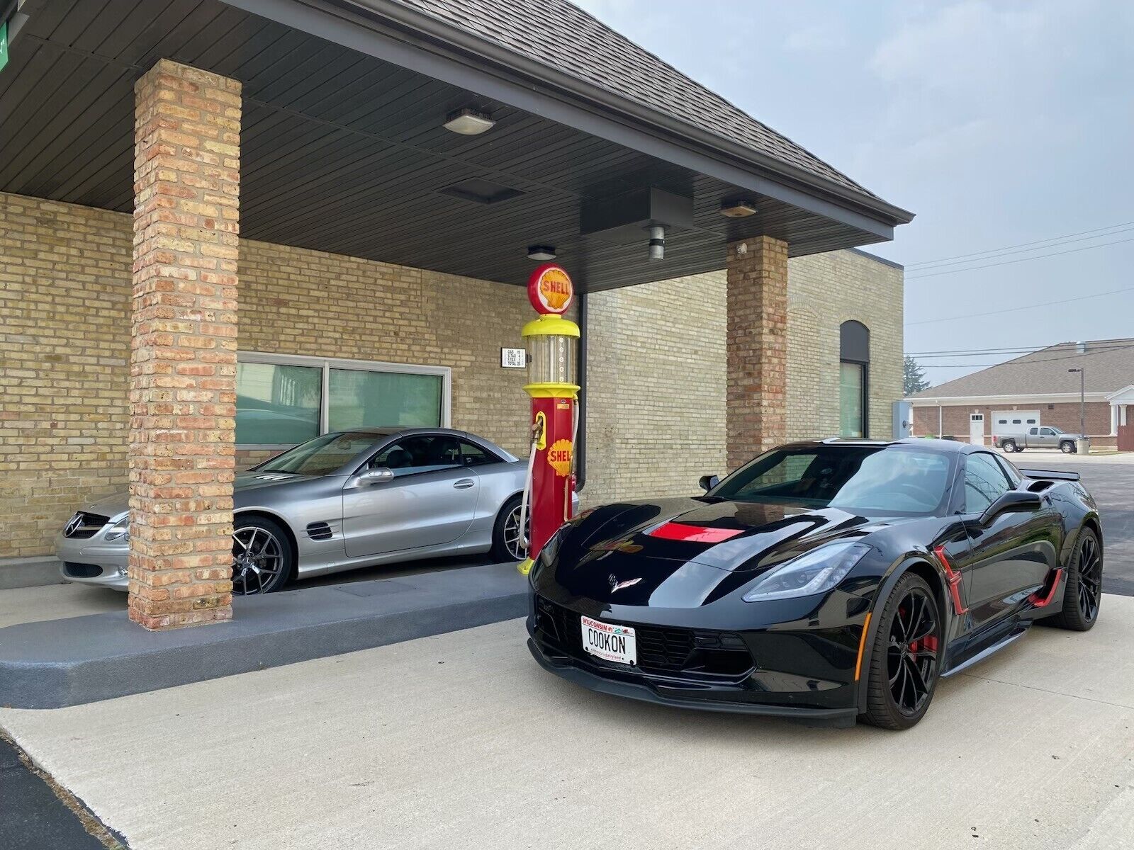 Une voiture de sport noire est garée devant un immeuble, sur lequel on peut voir l’enseigne d’une station-service Shell.