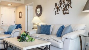 A living room with a white sofa, blue cushions, a coffee table with a plant, and wall art. - Fountain Hills Golf Condo | Heated Pool & Hot Tub (Fountain Hills)