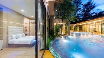 A modern bedroom with a large bed, a glass wall, and a view of a heated outdoor pool area.