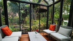 A conservatory with a wooden floor, two white cushioned sofas with orange pillows, a small table with a vase of a red flower, and a view of a lush garden through large glass panels. - TreeScape Resort Chiang Mai (Hang Dong)