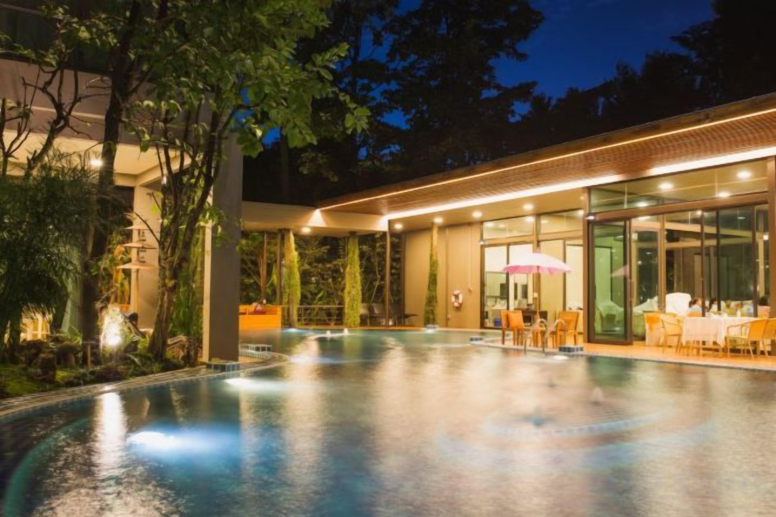 Un área de piscina al aire libre con una zona de estar cubierta y un edificio con cerramientos de vidrio.