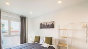 Ein Hotelzimmer mit Bett, einem fensterbelaubten Fenster, einem an der Wand befestigten Regal und einem Stadtbildgemälde.