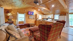 L’intérieur chaleureux d’un chalet avec un plafond en bois, un salon doté d’un canapé et de fauteuils à motifs, une cuisine avec des meubles en bois et un téléviseur fixé au mur.