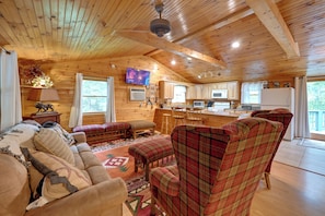 A cozy cabin interior with a wooden ceiling, a living room with a sofa and plaid chairs, a kitchen with wooden cabinets, and a television mounted on the wall. - 3 Mi to Lake Santeetlah! Rustic Apt w/ Decks (Robbinsville)