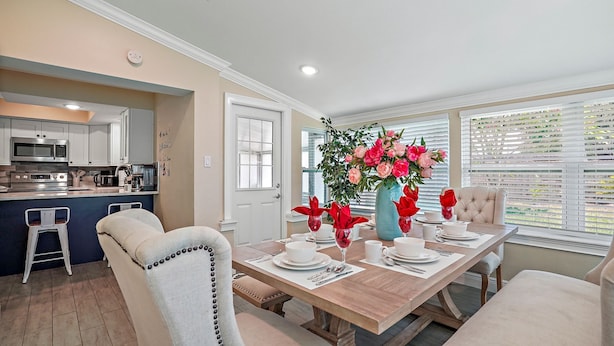 A dining area with a wooden table set for four, surrounded by cushioned chairs. A large vase with red flowers is on the table.