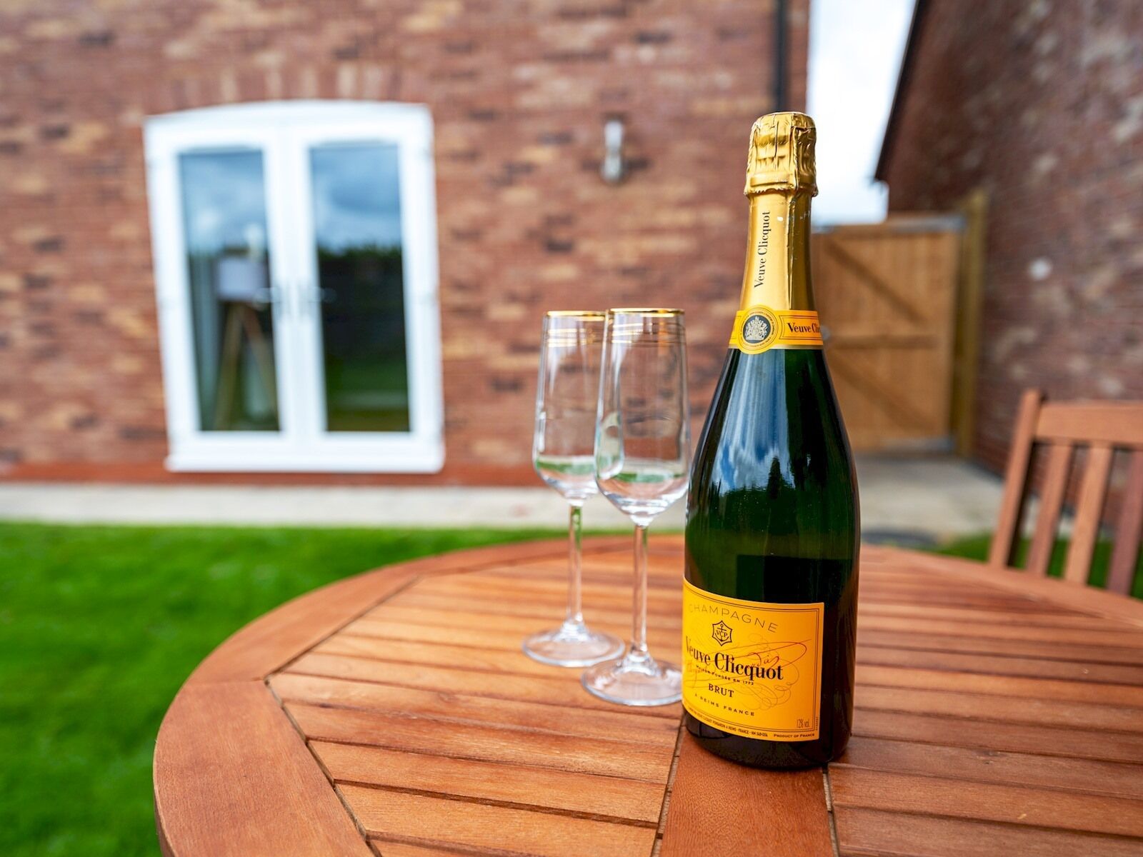 A bottle of champagne and two glasses on a wooden table outdoors.