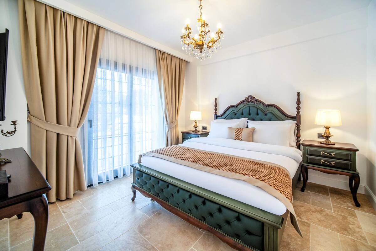 A spacious bedroom with a large bed, two bedside tables, a chandelier, and a view through a window with curtains.