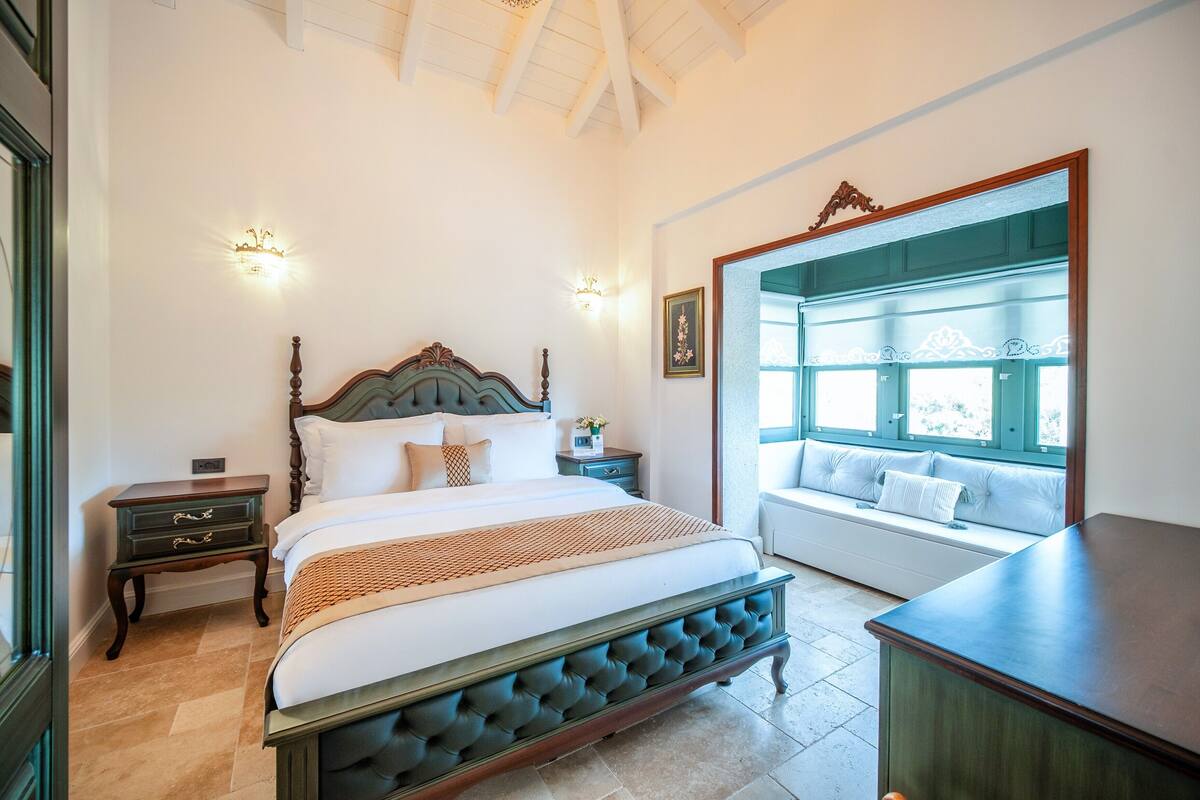 A bedroom with a large bed, a nightstand, a bench by the window, and a view of the outdoors.