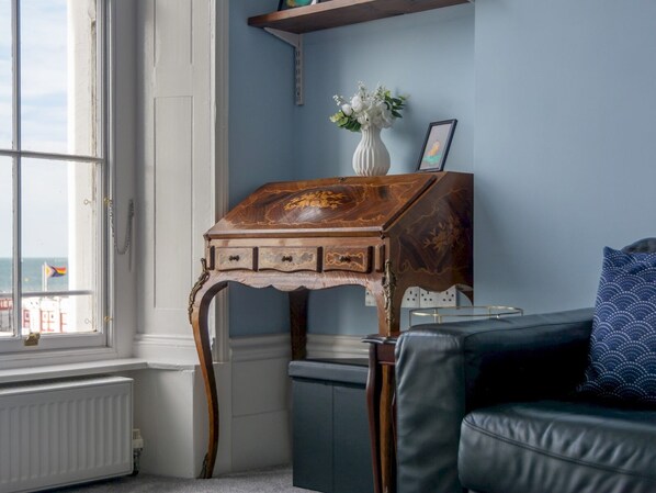 A room with a wooden desk, a vase with flowers, a framed picture, and a blue patterned pillow on a leather couch.