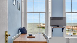 Ein Zimmer mit großem Fenster mit Blick auf das Wasser, einem Holztisch mit Stühlen, einem Fernseher auf einem Schrank und gerahmten Bildern an der Wand.