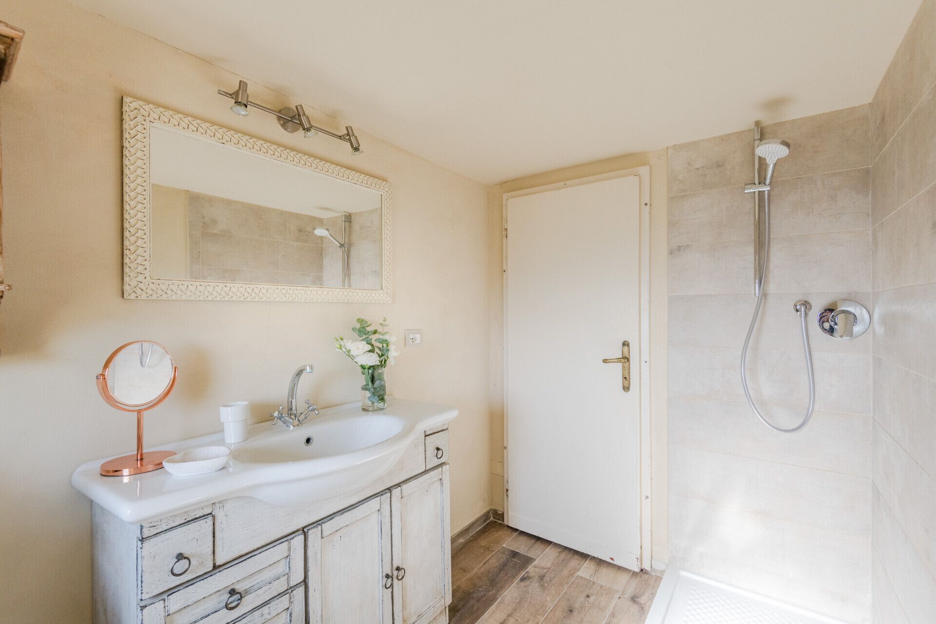 Une salle de bain avec un lavabo blanc, un miroir, un vase avec des fleurs, une douche et une porte.