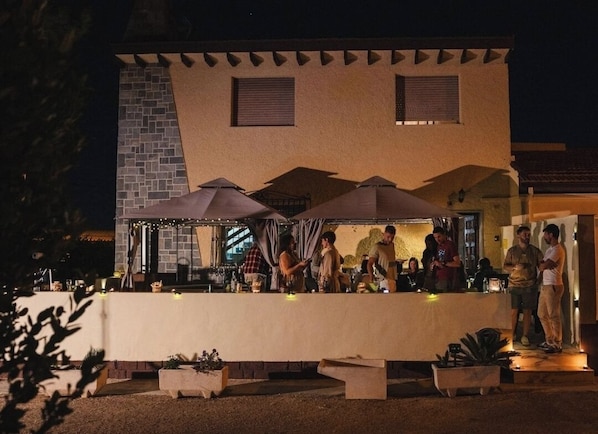 An outdoor bar area with a long counter, umbrellas, and a stone wall backdrop.
