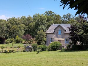 Une maison en pierre, dotée d’un toit à pignon, entourée d’une végétation luxuriante et d’arbres.