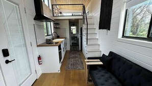 A compact, modern interior with a wooden floor, a black sofa, a small table, a ceiling fan, and a window with a view of trees. - Unit #5 - Modern Tiny Home in the heart of TX Wineries (Johnson City)