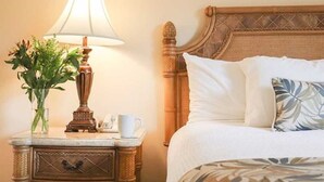 Ein ordentlich bezogenes Bett mit weißen Leinen und einem Zierkissen, ein Nachttisch mit Lampe, eine Vase mit Blumen und ein gerahmtes Bild an der Wand.
