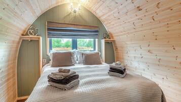 A cozy bedroom with a wooden sloped ceiling, a large bed with a quilted bedspread, two pillows, and a stack of folded towels.