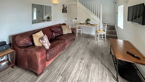 A living room with a brown leather sofa, a wooden dining table, and a staircase leading to another level. - 1 bedroom accommodation in Nether Wallop, Stockbridge (Nether Wallop, Stockbridge)