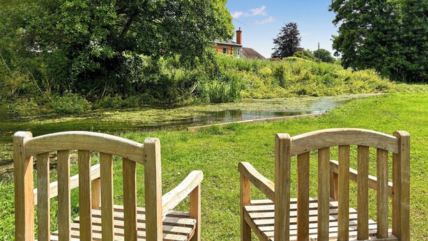 Two wooden chairs facing a river with a grassy bank and trees in the background. - 1 bedroom accommodation in Nether Wallop, Stockbridge (Nether Wallop, Stockbridge)