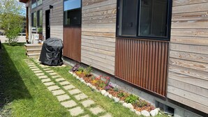 A modern house with a wooden exterior, large windows, and a stone pathway leading to a barbecue area. - Cozy 2 Bedroom by Teton Pass (Victor)